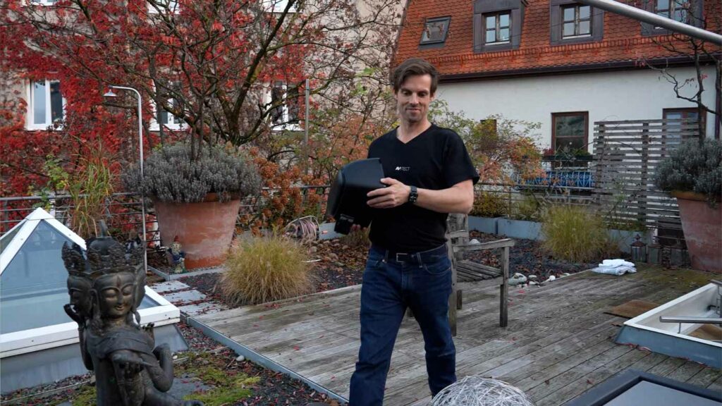Mann trägt Gerät auf Terrasse mit Herbstbepflanzung.