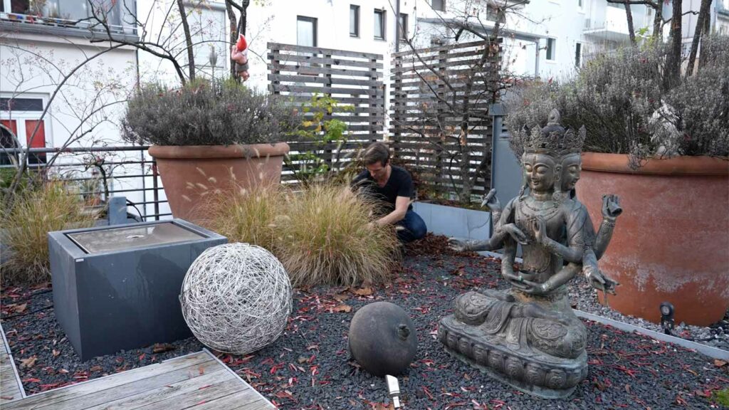 Mann pflegt Pflanzen auf bepflanztem Balkon mit Statue