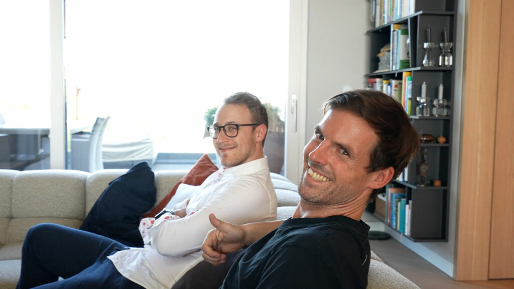 Zwei Männer lächeln in modernem Wohnzimmer.